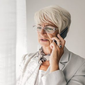 Woman speaking on a smartphone, wearing glasses and a light blazer. She stands near a window, appearing focused indoors.