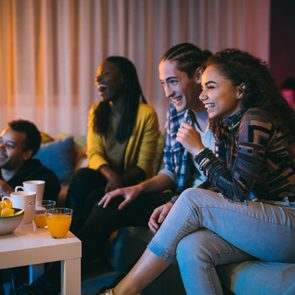 Four friends laugh while watching television, sitting on a sofa with snacks and drinks on a table in a cozy, dimly-lit living room.