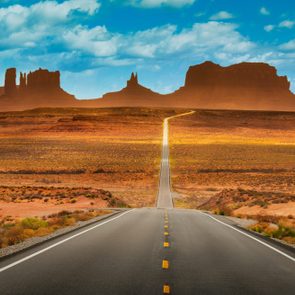 A straight road stretches toward distant mesas under a vibrant blue sky with white clouds, surrounded by desert landscape.