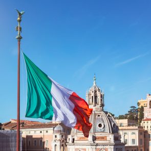 Italian flag waves on a pole, set against a clear blue sky and historic buildings in Rome.