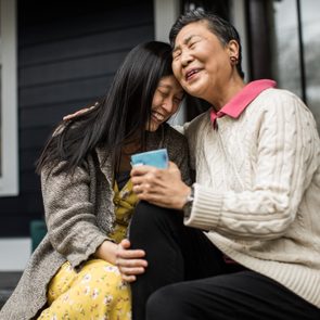 Two people sit closely, smiling and embracing, on a porch with dark siding. One holds a blue mug.