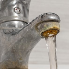 A metal faucet with water flowing, against a plain light background. The surface of the faucet appears worn and slightly corroded.