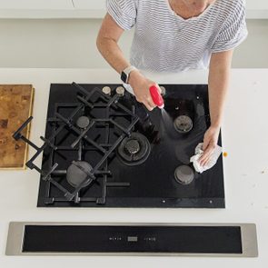 Person cleans a stovetop, spraying cleaner and wiping with a cloth in a modern kitchen. A wooden cutting board is nearby.