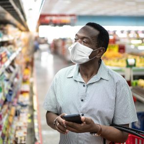 A person wearing a mask holds a smartphone, browsing grocery items in a supermarket aisle.