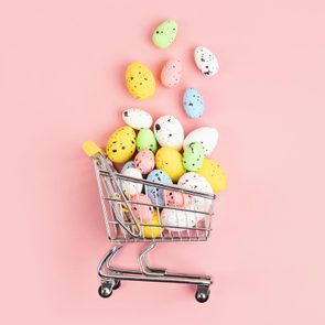 Colorful speckled eggs spill from a small shopping cart, set against a light pink background.