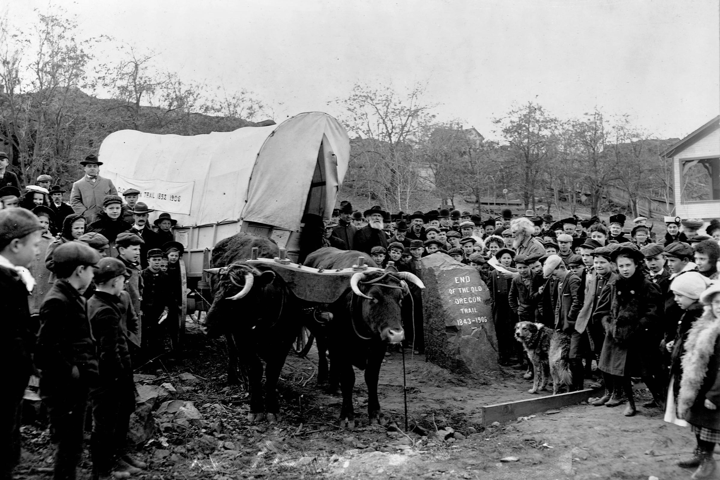 A crowd of the descendants of Oregon Trail settlers gathers together in The Dalles, Oregon, for a celebration at the inscribed stone that marks the trail's end. A covered wagon pulled by a team of oxen completes the occasion.