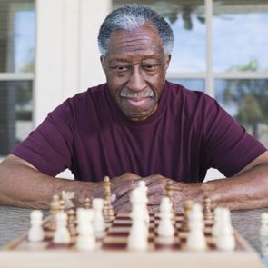 Man thoughtfully plays chess, wearing a maroon shirt, seated outdoors near a large window.
