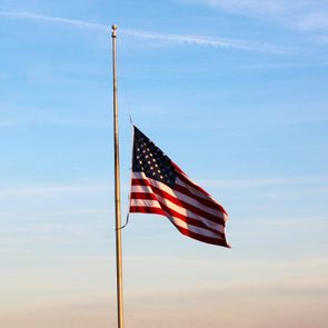 Flag flies at half-mast on a flagpole, against a clear blue sky background.