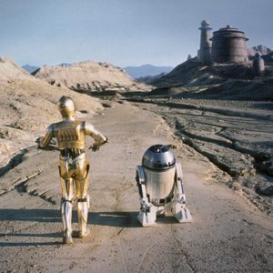 Two robots, one gold and humanoid, the other small and dome-shaped, travel through a barren, desert landscape toward distant, futuristic buildings.