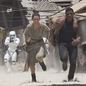 Two people run urgently through a sandy, chaotic marketplace, pursued by white-armored figures with weapons aimed.