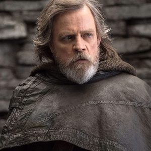 Bearded man wearing a cloak stands, looking to the side, against a textured stone wall background.