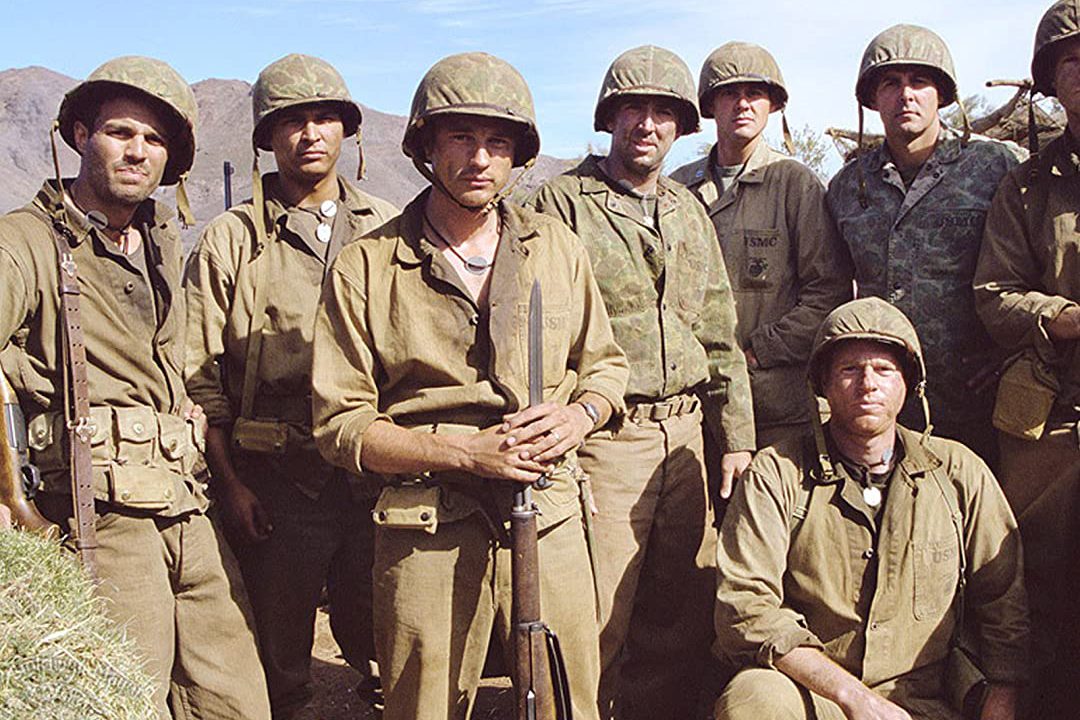 Soldiers stand together, wearing World War II military uniforms and helmets, in a mountainous landscape under a clear sky.