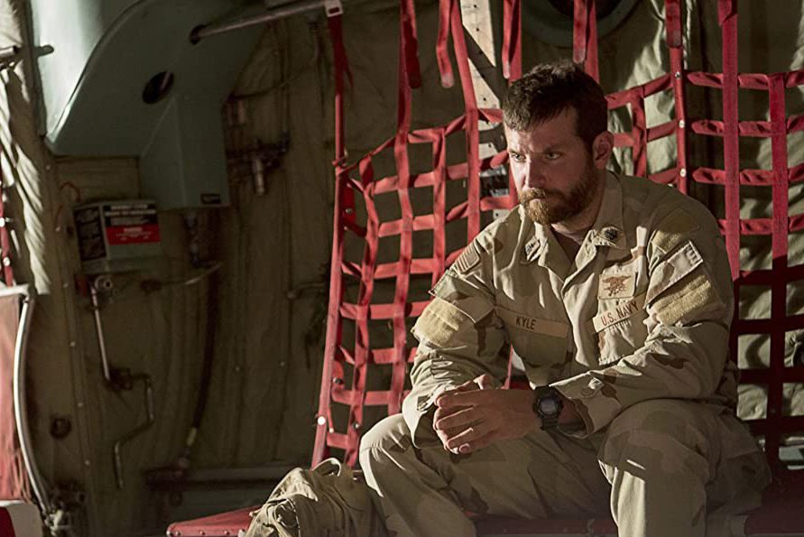 A soldier sits pensively in military fatigues inside an aircraft, surrounded by netted red seats.