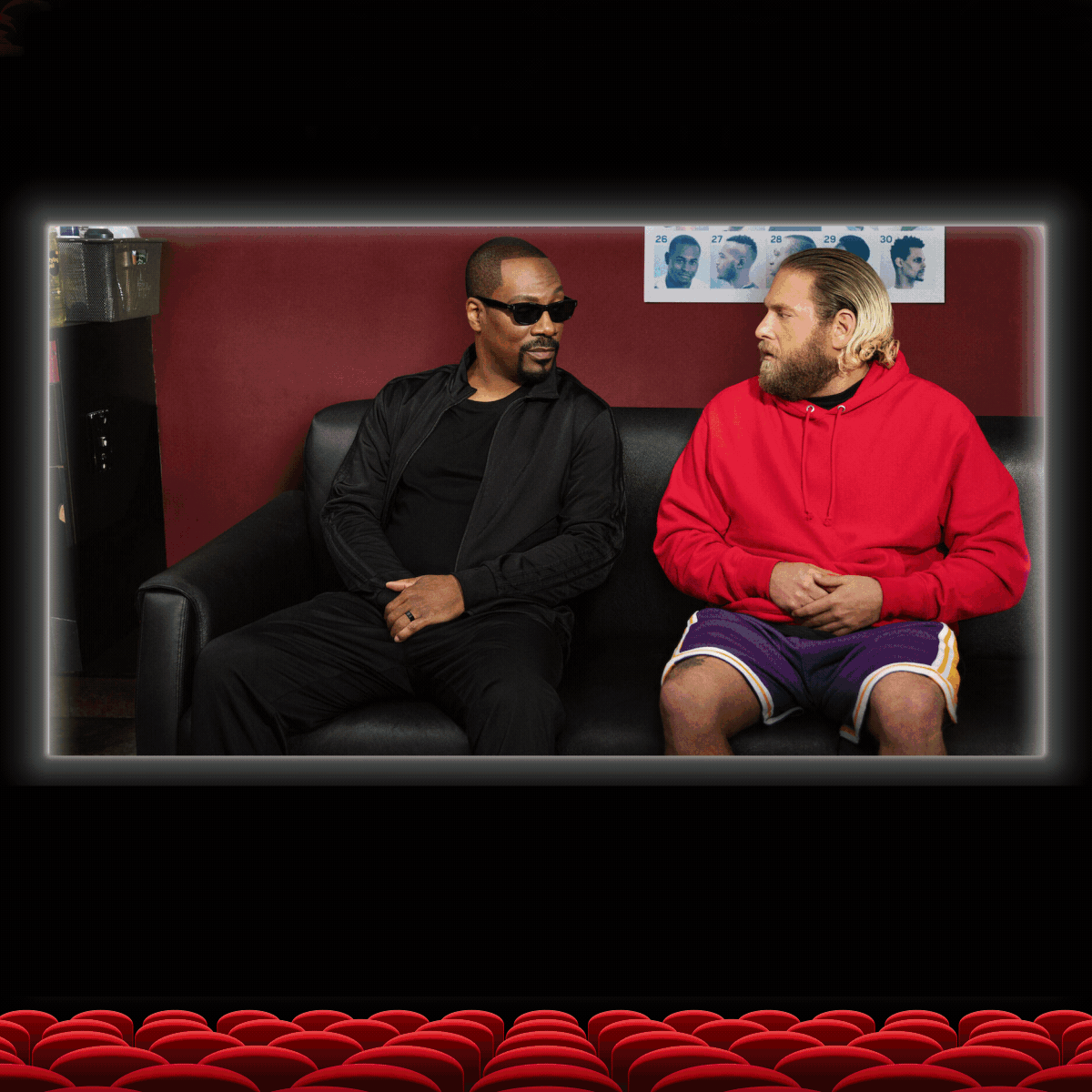 Two men sitting on a black couch, one in black outfit and sunglasses, the other in a red hoodie, with salon photos behind them.