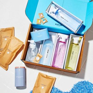 Cleaning product set displayed in an open box, featuring colorful bottles and packets, on a bright surface with blue rug nearby. Text includes brand 