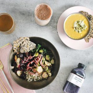 A bowl filled with roasted vegetables and seeds sits on a gray table with drinks, a soup bowl, and a purple juice bottle nearby.