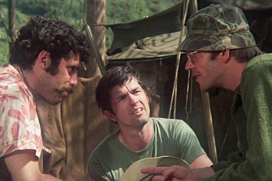 Three men converse intensely, one wearing a camo hat, outdoors in front of army tents.