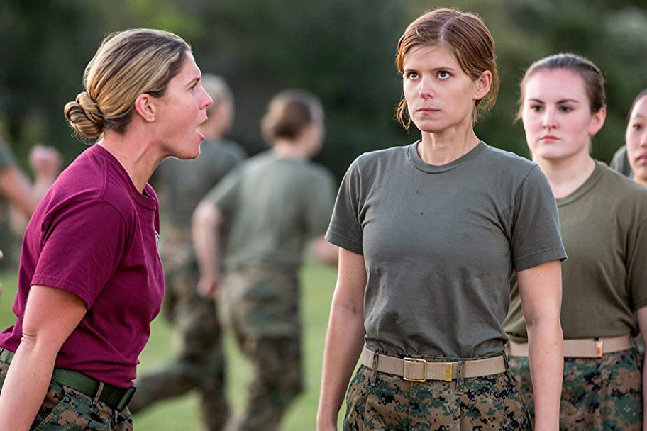 Instructor shouts at trainee, standing at attention, amidst a military training field with others in uniform.