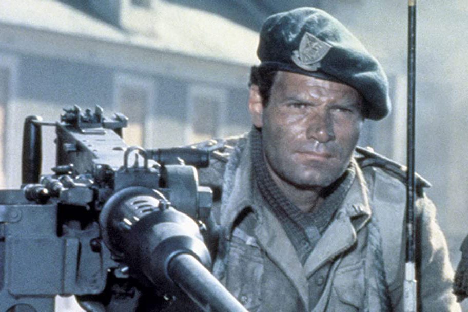 A soldier in uniform, wearing a beret, mans a large machine gun outdoors, beneath a bright sky, near a building.
