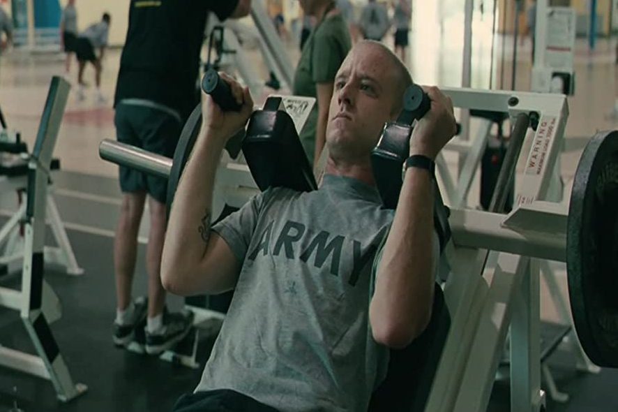 Man lifts dumbbells on a gym machine, wearing a "ARMY" shirt, surrounded by other gym-goers and equipment.