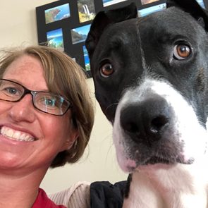 A smiling woman and a black-and-white dog sit indoors, with a photo collage on the wall behind them.