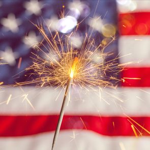 A lit sparkler emits sparks against a blurred background of the American flag's stars and stripes.