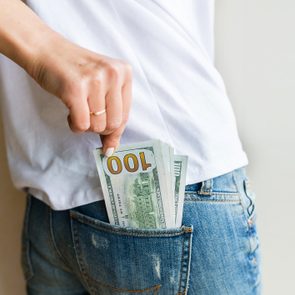 Hand placing several 100-dollar bills into a back denim pocket, against a plain wall.