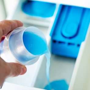 A hand pours blue liquid detergent into a washing machine drawer compartment, set against a blurred blue and white background.