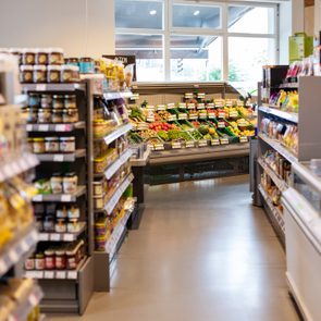 Store shelves display various packaged goods beside a fresh produce section. Brightly lit interior with large windows at the far end.