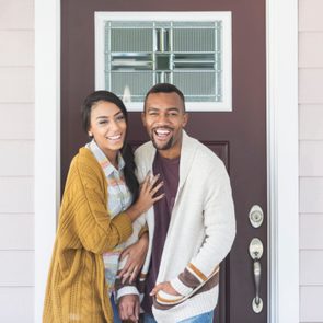 A smiling couple standing in front of a brown door with decorative glass, surrounded by light-colored siding.