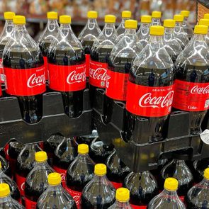 Coca-Cola bottles with yellow caps are arranged on black crates in a store display, surrounded by various other products.