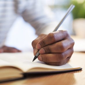 Hand writes in a notebook on a wooden table, the background slightly blurred.