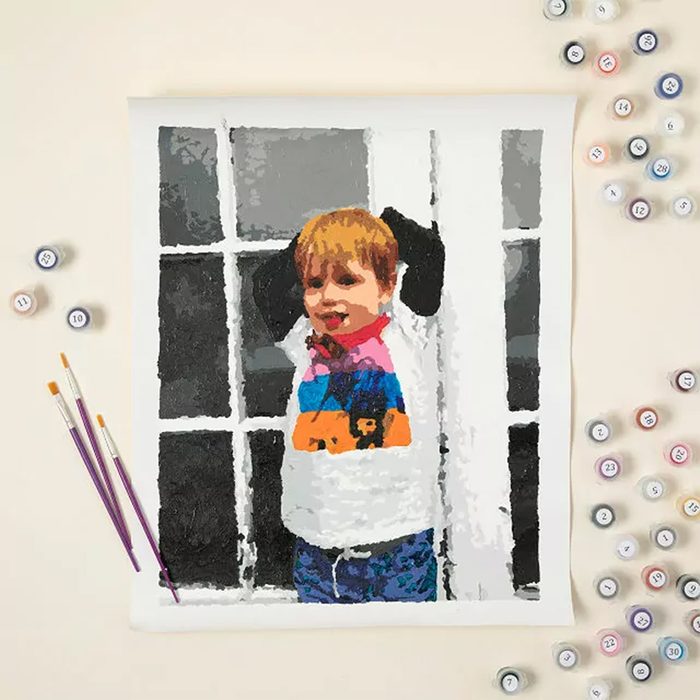 Painting partially completed on canvas, depicting a child with raised arms, set beside paint pots and brushes, on a light surface.