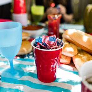 Red cup holds small American flags on a festive table with food and drinks, including buns and beverages, under sunlight.