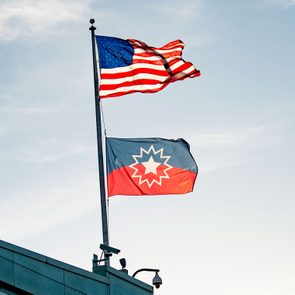 Two flags wave atop a building; the American flag and a blue, red flag with a starburst. Clear sky in the background.