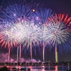 Fireworks explode in vibrant red, white, and blue colors above a calm river, with silhouetted trees and city lights in the background.