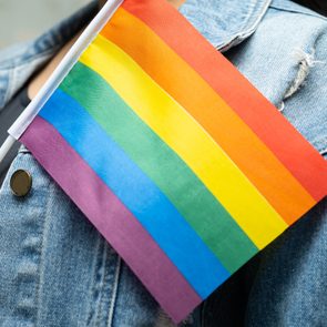 A rainbow flag is held by a person wearing a denim jacket, symbolizing LGBTQ+ pride in an outdoor setting.