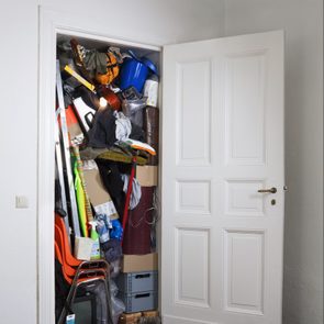 Overflowing closet holds various items, clutter spilling out, surrounded by a plain white room.