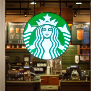 Lit Starbucks logo hangs in a coffee shop window, surrounded by menu boards and a counter with various drinks and equipment.