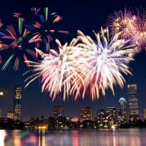 Fireworks explode vibrantly over a city skyline at night, reflecting on a calm river below.