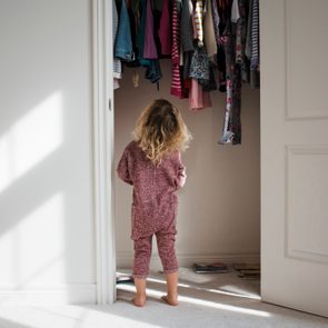 Child stands facing a closet filled with hanging clothes, in a sunlit room with white walls and carpet.
