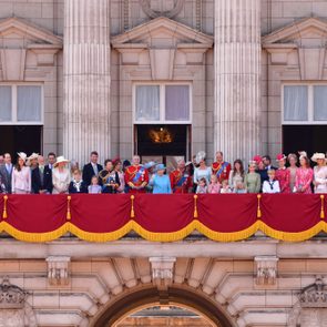A large group of people stand on a balcony adorned with red and gold fabric, in front of a grand, historic building facade.