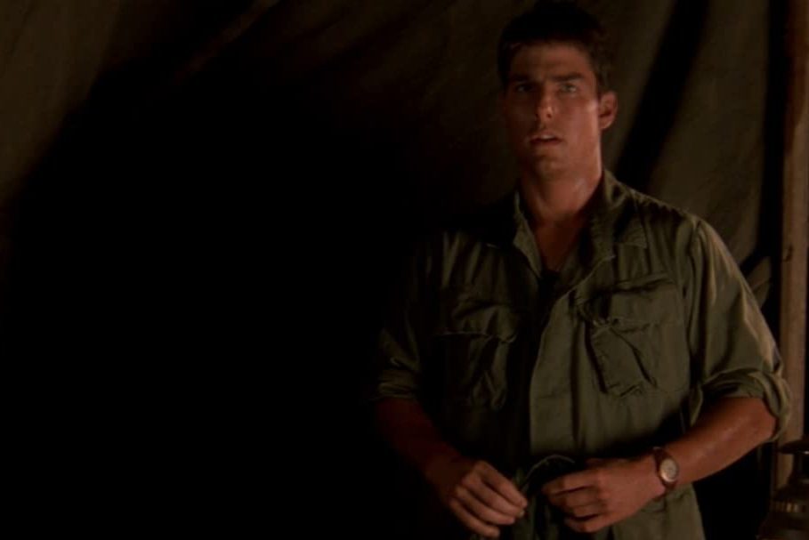 A person, standing, adjusts their shirt in a dimly lit tent wearing a green military-style outfit.