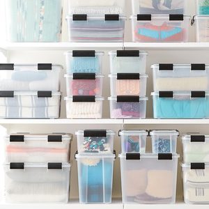 Clear plastic storage bins, filled with neatly folded clothes, stack on white shelves in a well-organized, bright room.