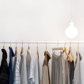 Clothes hang on wooden hangers along a white rod under a glowing light bulb, against a plain white wall.