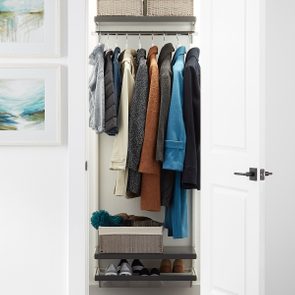 Coats hang neatly in an organized closet with shelves holding shoes and baskets, inside a white-walled room with abstract paintings.