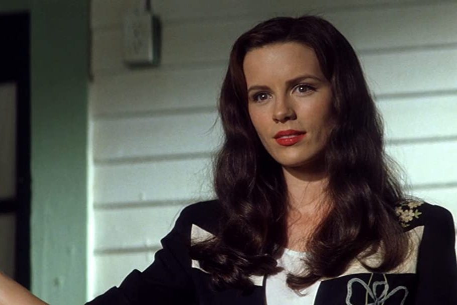 Woman stands indoors, gazing calmly, wearing a black and white patterned jacket, with white paneled walls in the background.