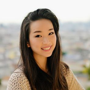 A person with long hair smiles warmly, wearing a knit sweater, standing in front of an out-of-focus urban background.