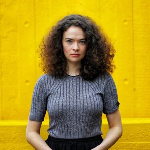 Person stands still against a bright yellow wall, wearing a grey ribbed shirt, with curly hair and a serious expression.
