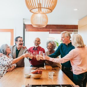 Group clinks glasses, smiling around a kitchen island with bread and grapes, under elegant wooden pendant lights.